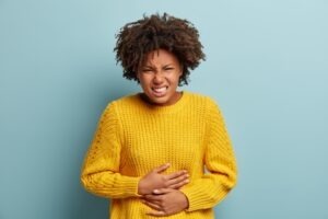 Displeased dark skinned woman suffers from stomachache, period cramps, keeps hands on belly, wears yellow knitted sweater, smirks face, stands against blue background. People and sickness concept Displeased dark skinned woman suffers from stomachache, period cramps, keeps hands on belly, wears yellow knitted sweater, smirks face, stands against blue background. People and sickness concept